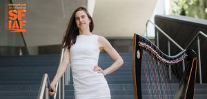 A woman in a white dress posing next to a harp at an arts festival event.
