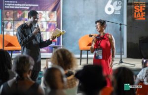 An author reads from a book at a literature event while an attentive audience listens, against the backdrop of a festival banner.