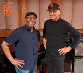 Two men smiling and posing together in a studio with musical equipment in the background.