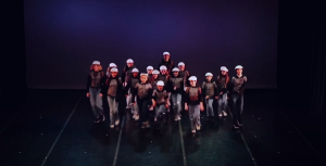 A group of dancers in white hats and grey outfits performing a choreographed routine on stage.