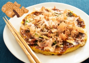 Okonomiyaki (japanese savory pancake) topped with bonito flakes, with chopsticks and a decorative fish-shaped element on the side.