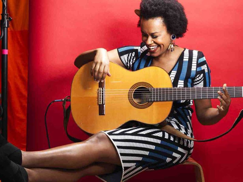A woman sitting and laughing while holding a guitar against a red background.