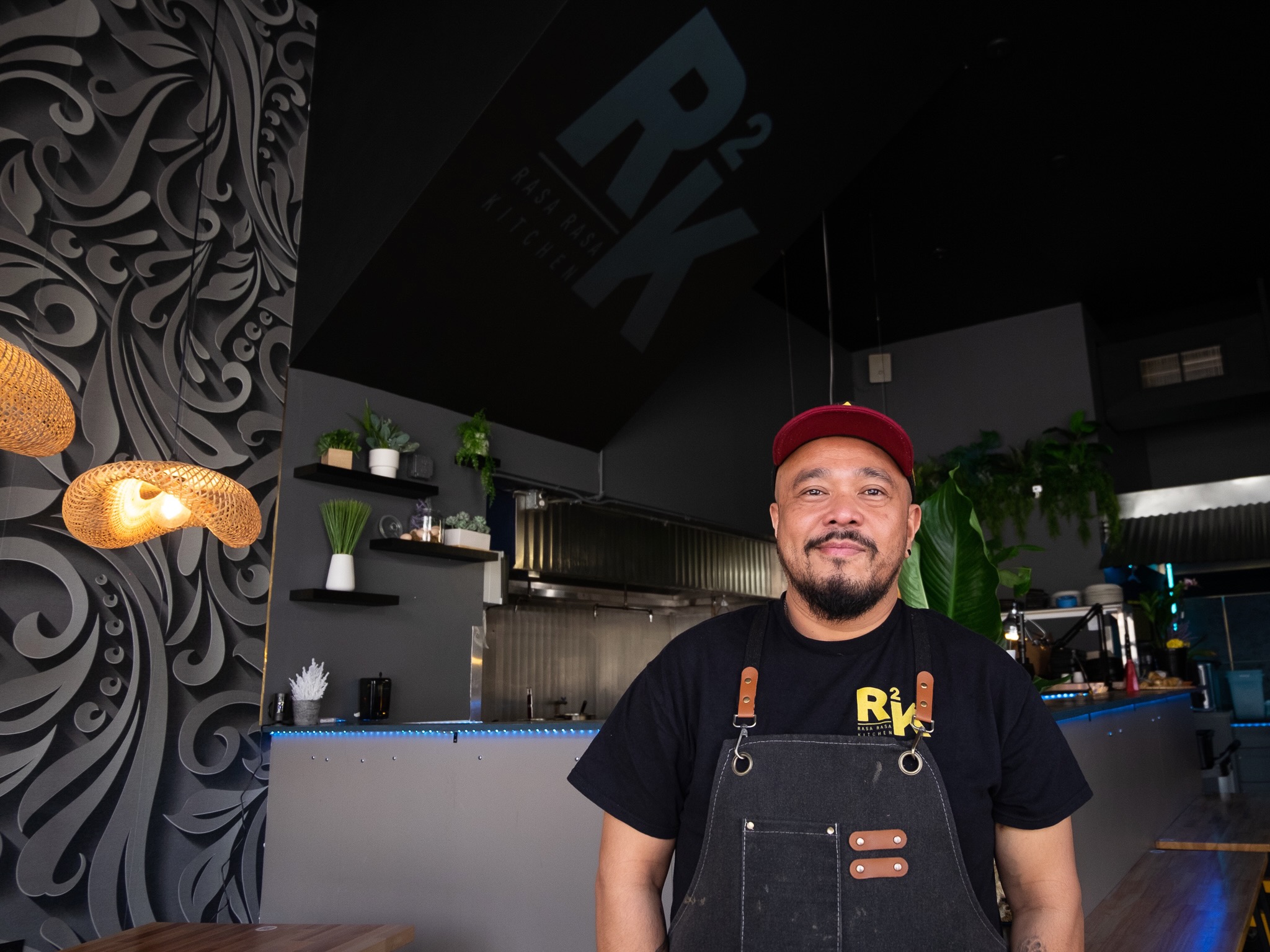 A chef in a black apron stands proudly in a modern restaurant kitchen.
