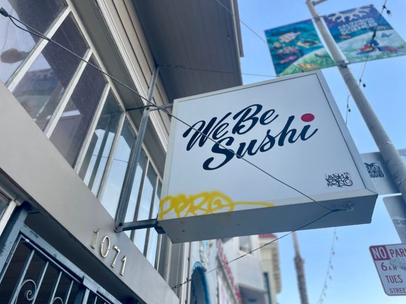Signboard reading "We Be Sushi" mounted on a building's facade with graffiti on the side.