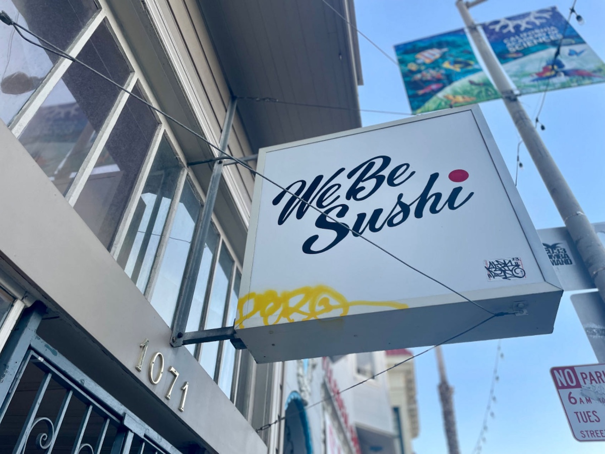 Signboard reading "We Be Sushi" mounted on a building's facade with graffiti on the side.