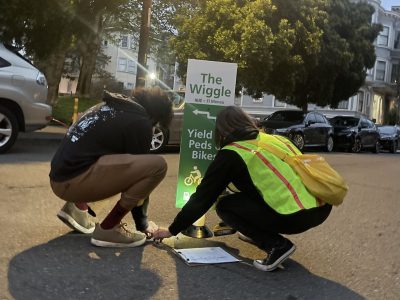 Two people, one in a reflective vest, doing something on a street.