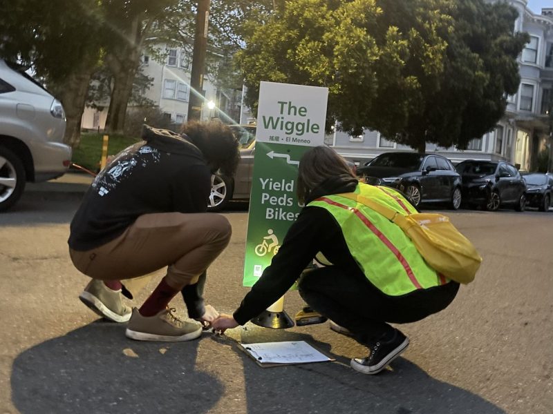 Two people, one in a reflective vest, doing something on a street.