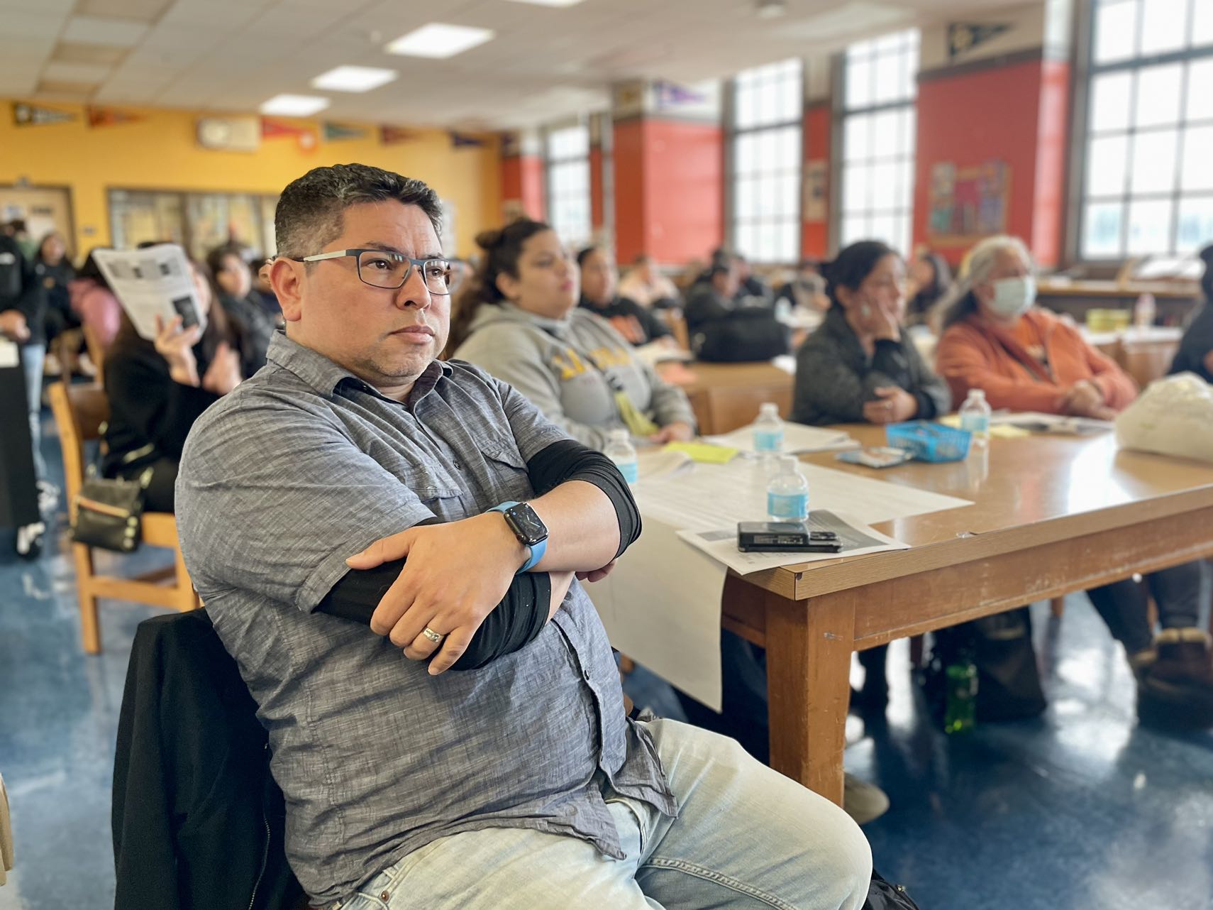 A man sitting with crossed arms at a Mission High SFUSD meeting, with other attendees in the background. meeting on school closures.