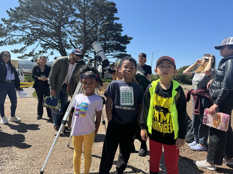A group of people with children standing outdoors during a daytime event, with a telescope set up for astronomical observation.