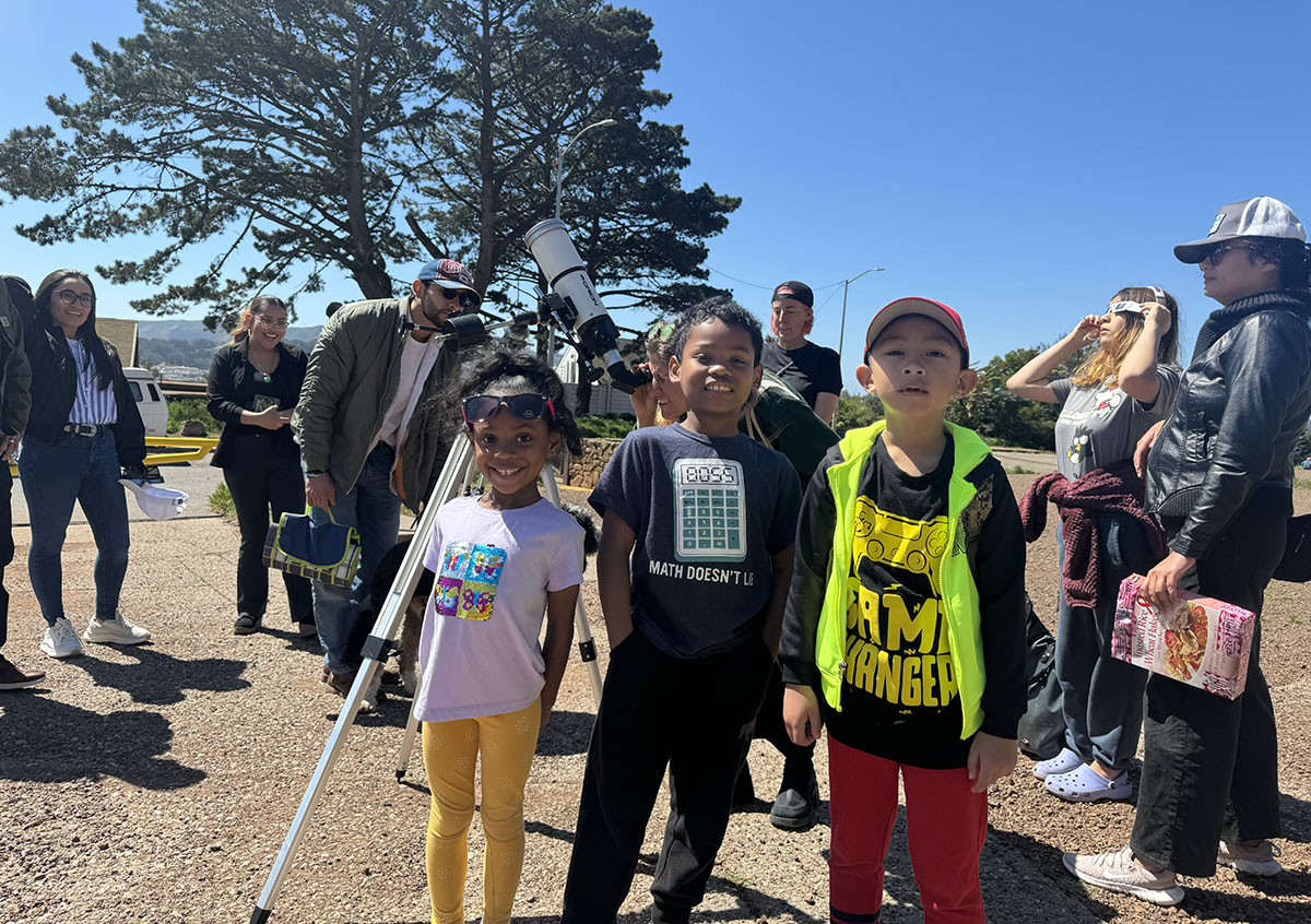 A group of people with children standing outdoors during a daytime event, with a telescope set up for astronomical observation.