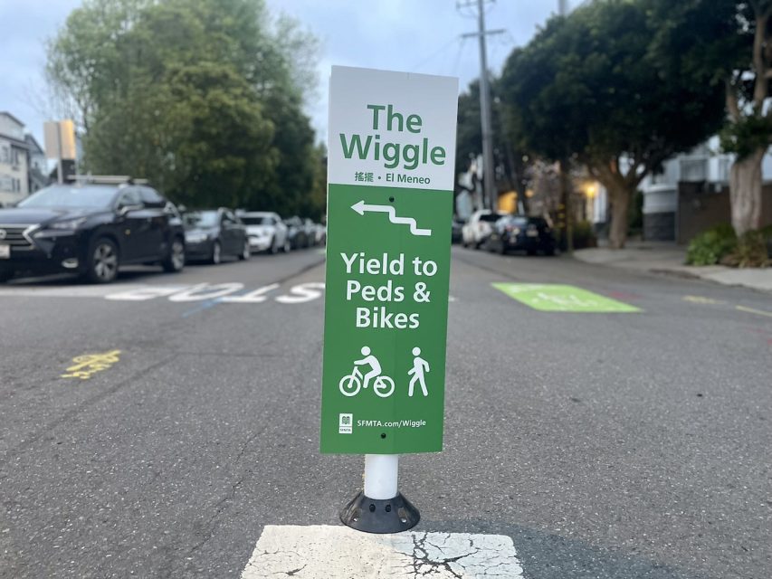 Green traffic sign instructing to yield to pedestrians and bikes, positioned on a street with cars and houses in the background.