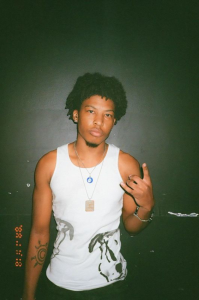 A young man in a white tank top with tattoos poses against a dark background, making a peace sign with his hand.