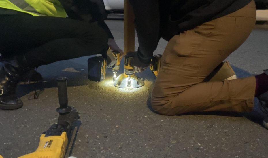 Two people installing a slow street bollard at night, illuminated by a flashlight, with tools nearby on the ground.