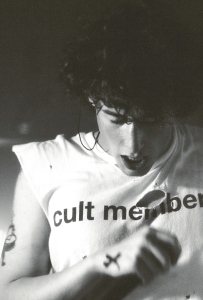 A black and white image of a young person in a "cult member" t-shirt intensely looking at a tattoo on their arm.