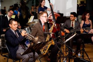 Jazz ensemble performing with a saxophonist and trumpeter in the foreground.