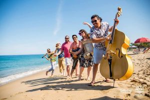 A group of musicians joyfully playing instruments and dancing on a sunny beach, featuring a man with a double bass in the foreground.