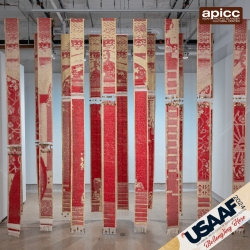Art installation featuring a series of vertical textile banners with intricate red patterns hanging from a ceiling in a gallery space.