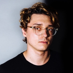 Portrait of a young man with curly hair, wearing glasses and a black shirt, looking directly at the camera against a dark background.