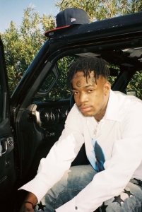 A young man with tattoos sits on a car's running board, wearing a white shirt and camouflage pants, with a baseball cap resting on the car roof above him.