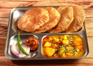 Three puri breads on a stainless steel tray with a side of potato curry, onion slices, and spicy chutney.
