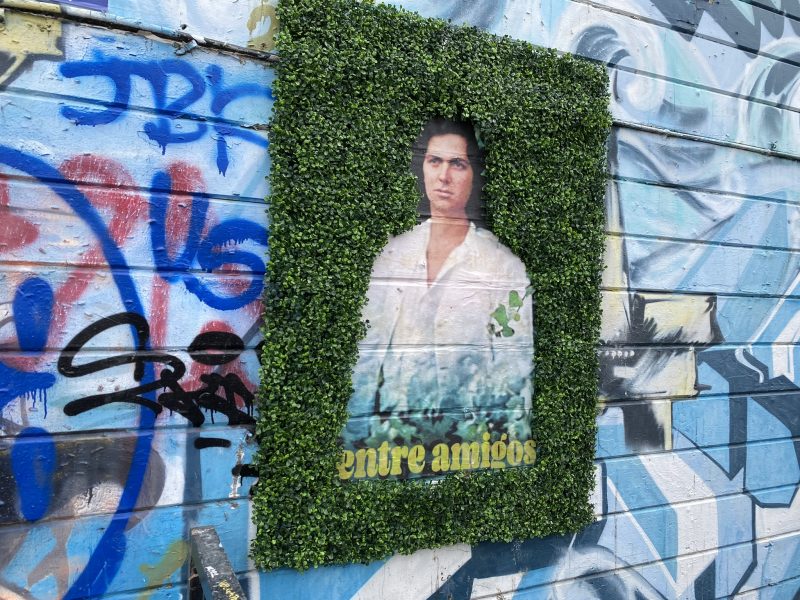 A poster on a graffitied wall with green background and a woman's picture in the center with Spanish words "entre amigos" under the woman's photo.