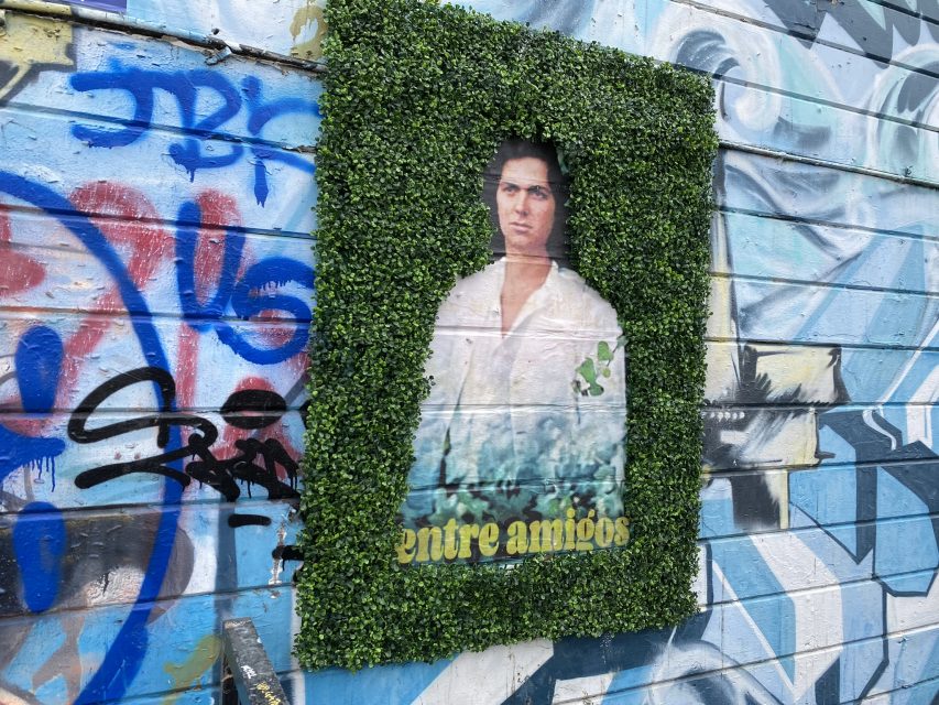 A poster on a graffitied wall with green background and a woman's picture in the center with Spanish words "entre amigos" under the woman's photo.