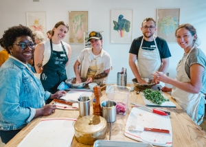 Five adults smiling at the camera in a cooking class, wearing aprons and surrounded by cooking utensils and ingredients on a kitchen counter.