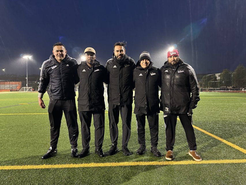 Five men standing together on a soccer field at night, wearing black jackets and smiles; it's raining lightly.