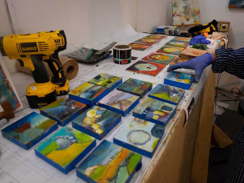 An artist's worktable covered in colorful, abstract paintings, with various art supplies and a power tool nearby.