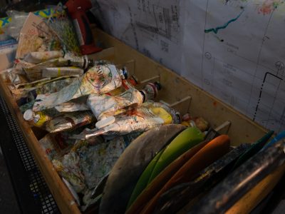 A cluttered artist's tray filled with used paint tubes, brushes, and palettes, set against blueprints in the background.