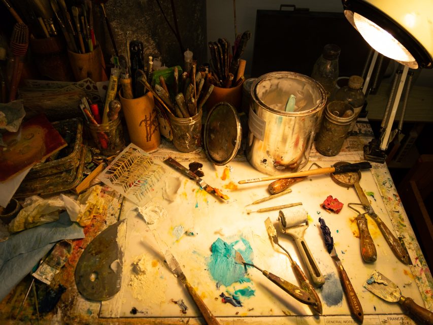 A cluttered artist's desk with scattered painting tools, brushes, and vibrant paint smears under a desk lamp.