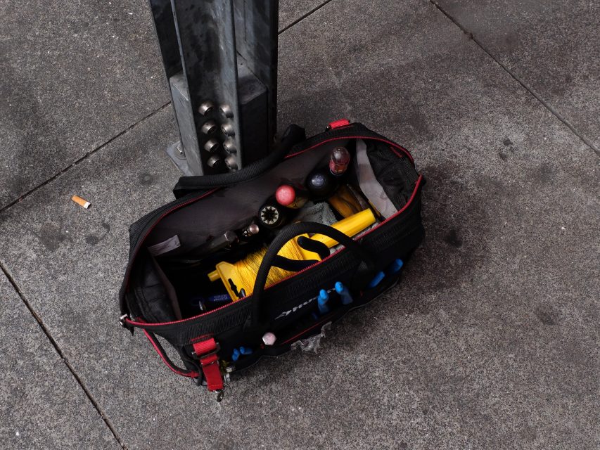 An open red and black equipment bag on a gray sidewalk, filled with various juggling balls and yellow pins.