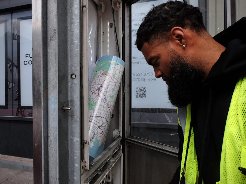A man in a neon vest examines a city map displayed inside a public transportation shelter.