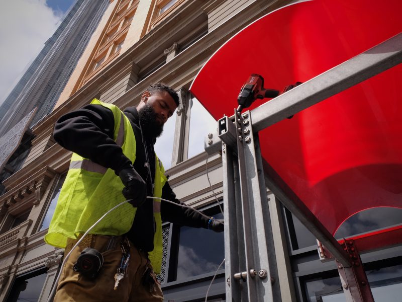 A worker in a reflective vest and tool belt adjusts equipment on a large red structure on a city street.