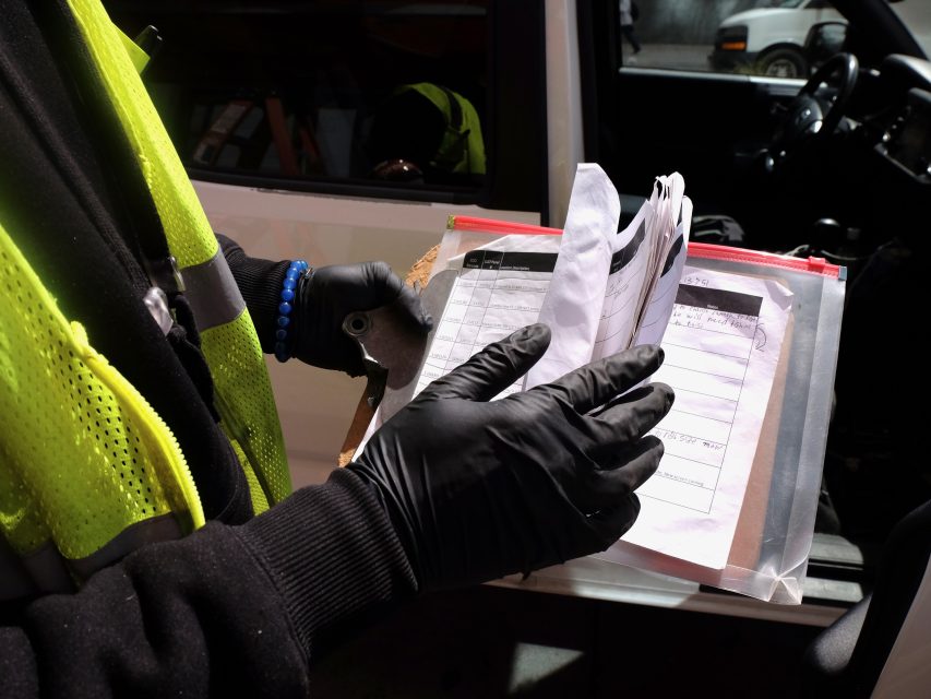 A worker in a reflective vest and gloves reviews paperwork in a clipboard, in a sunlit outdoor setting.