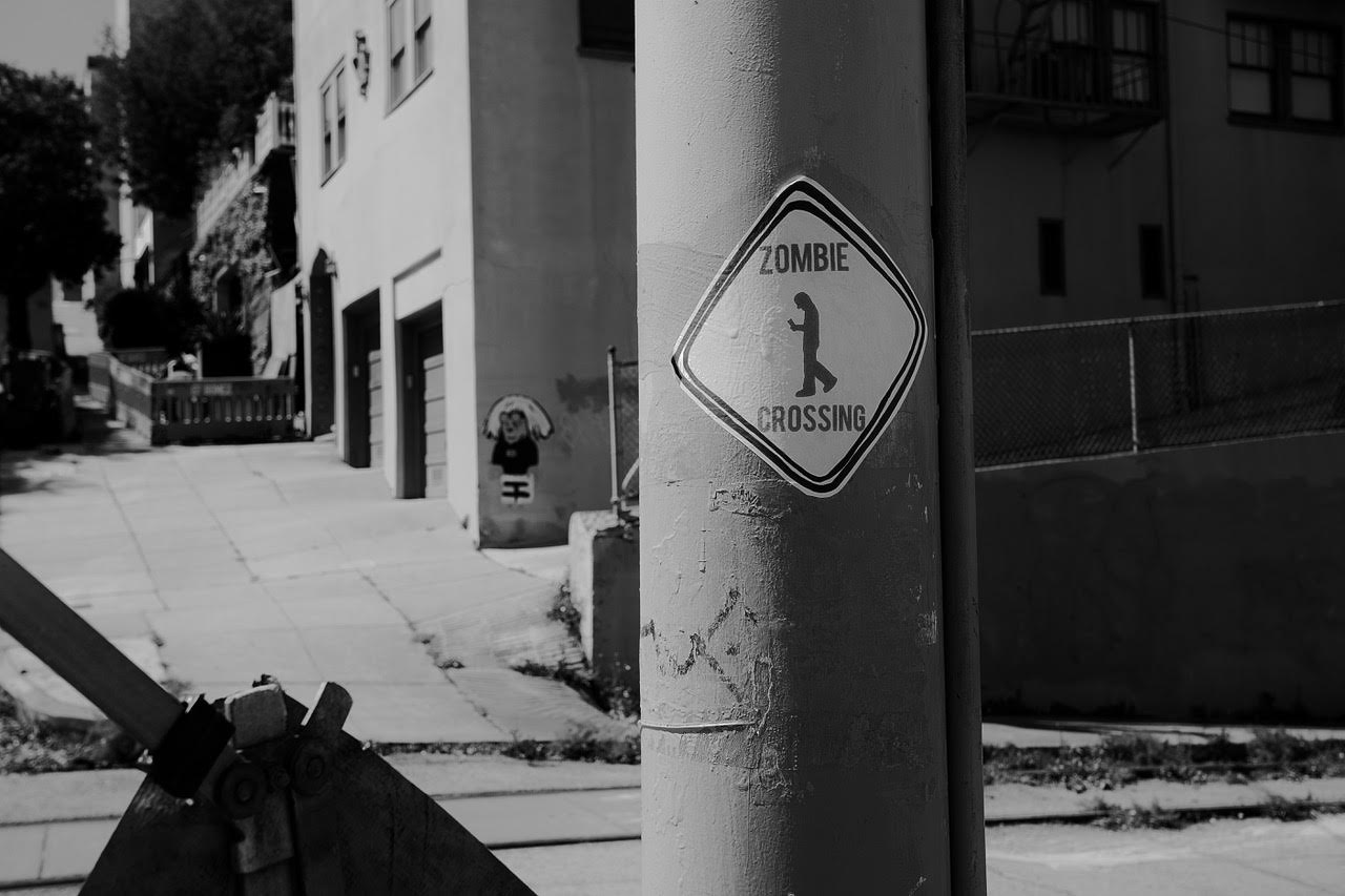 A black and white photo showing a street sign with "zombie crossing" and a graphic of a pedestrian walking with head down looking at their phone , on an urban street.