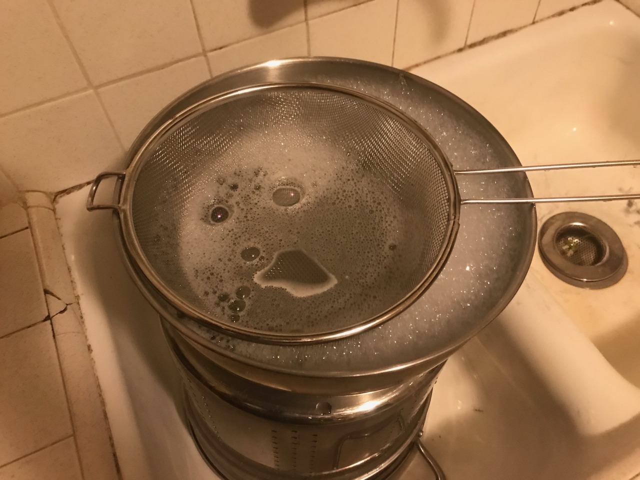 A pot filled with soap water with bubbles on the surface sitting in the sink in dim lighting. A strainer is rested on the pot with bubbles coming through.