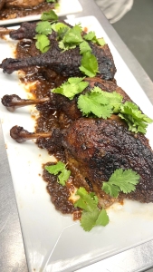 Four roasted chicken drumsticks covered in a dark glaze and garnished with cilantro leaves on a white rectangular plate.