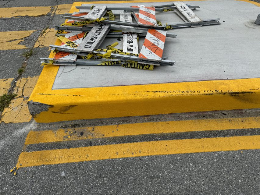 Pile of damaged and disassembled road signs stacked on a concrete median with bright yellow and black markings.