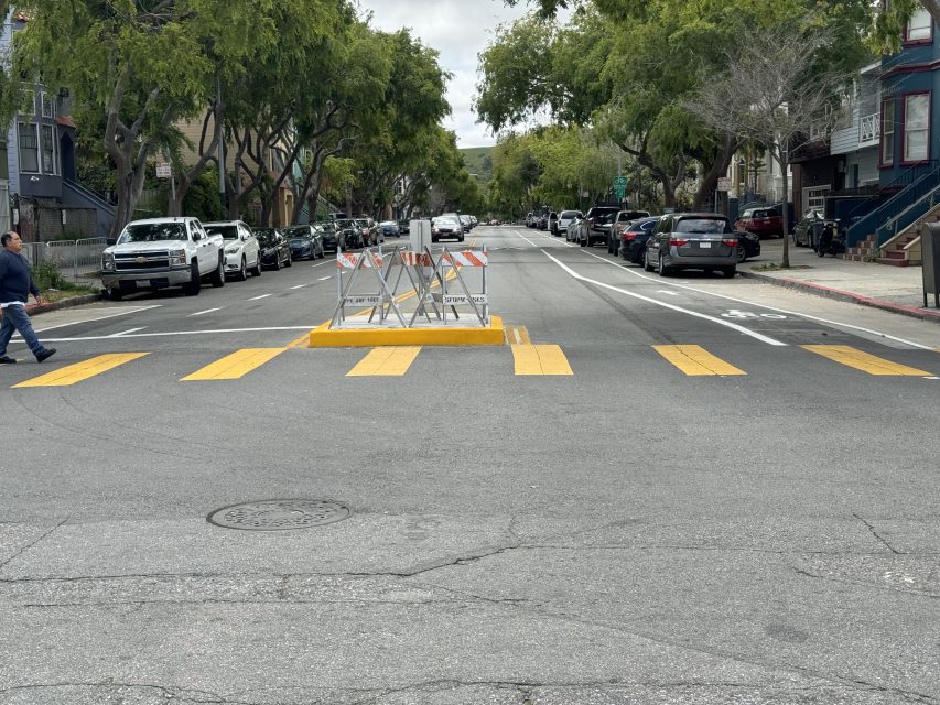 A street barrier with yellow markings blocks a road, surrounded by parked cars and trees, with a pedestrian on the left.