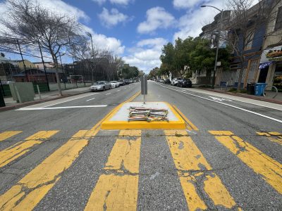 A city street view showing a yellow pedestrian crossing with a median strip containing newspaper stands, flanked by cars and buildings under a cloudy sky.