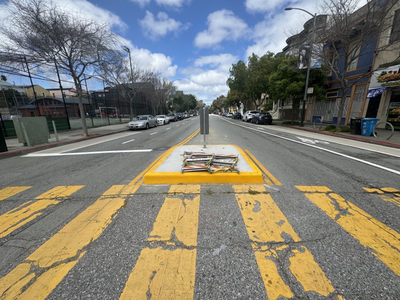 A city street view showing a yellow pedestrian crossing with a median strip containing newspaper stands, flanked by cars and buildings under a cloudy sky.