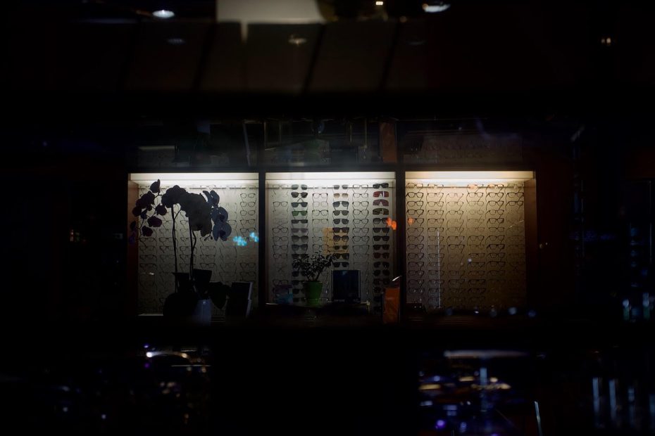 In the dark, a window of the shop is lit up. In the window are shelves with glasses displayed on them.