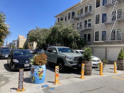 Cars parked on a sunny street with traffic cones and decorated utility boxes, residential buildings in the background.