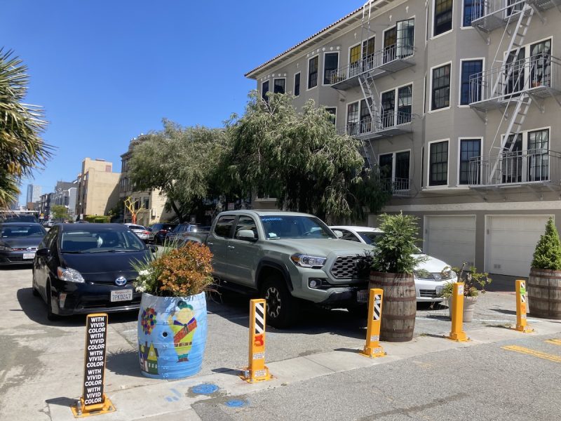 Cars parked on a sunny street with traffic cones and decorated utility boxes, residential buildings in the background.