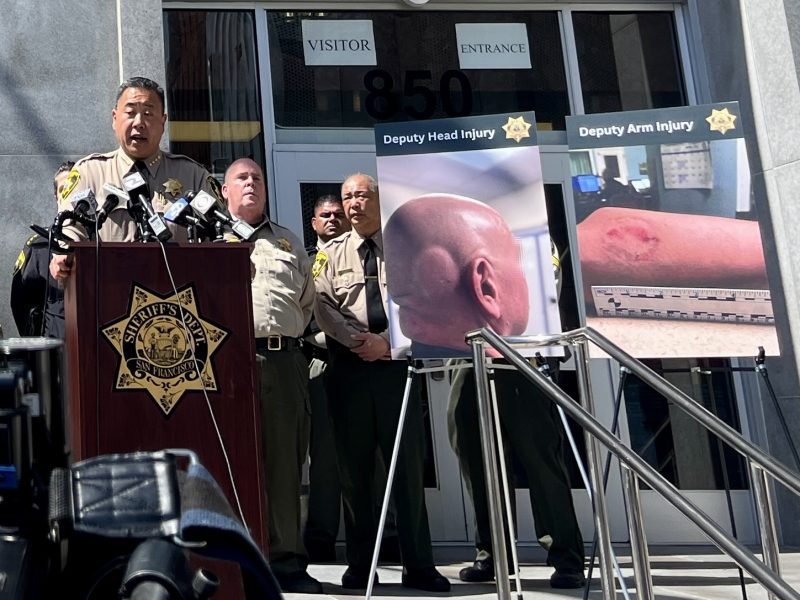 Sheriff giving a press conference outside a building, with images showing injuries displayed next to him.