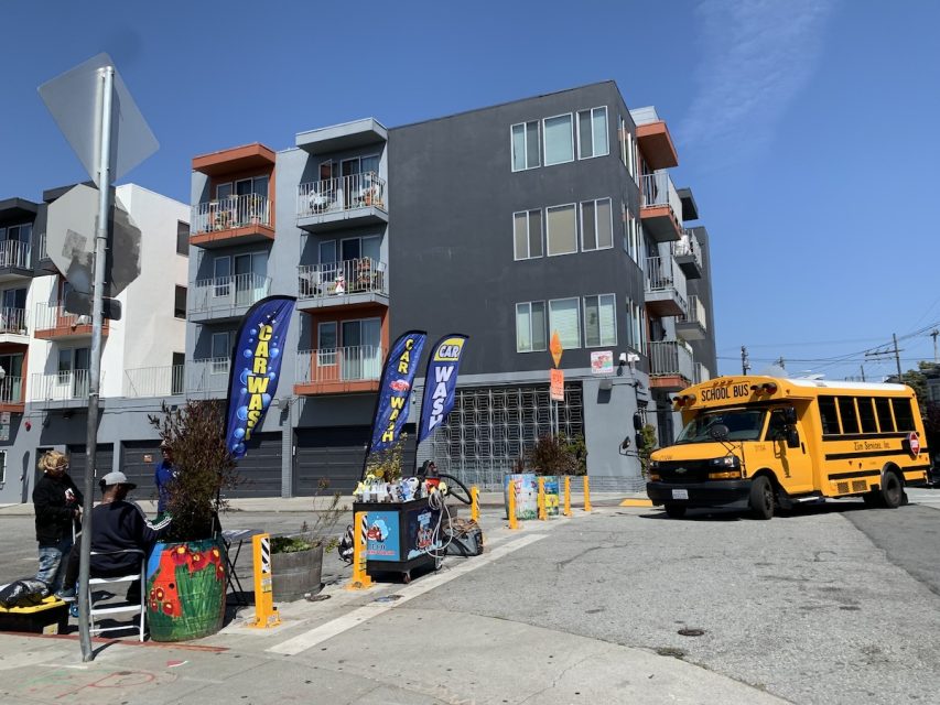 Sunny street view showing a modern gray apartment building, a yellow school bus, and two people with bicycles near colorful flags and planters.