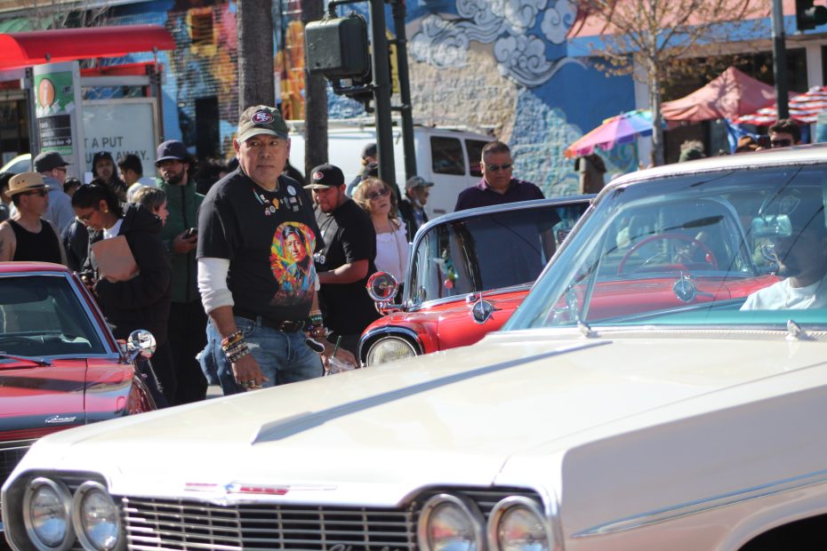 An attendee wearing a Cesar Chavez t-shirt watches the lowriders drive by during the Selena lowrider tribute