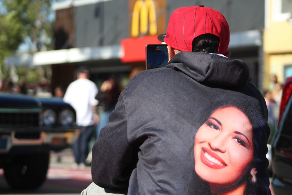A man with a Selena hoodies takes a picture of lowriders cruising by on Mission Street during the Selena lowrider tribute.