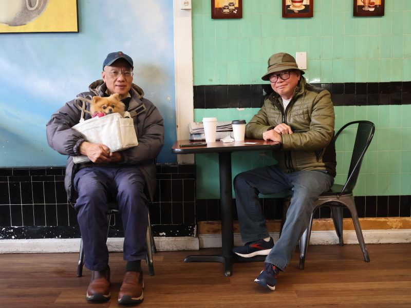 两名中国男子分别坐在咖啡馆的两张桌子旁，其中一人怀里抱着一只小狗。